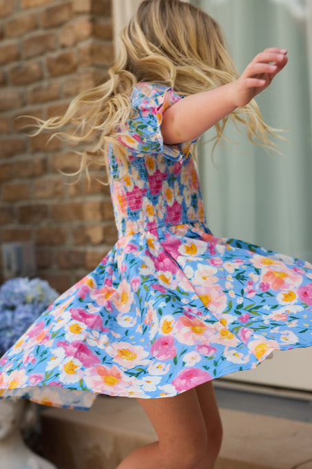A young girl with blonde hair twirls outdoors in the Ivy City Co Mini Teagan Dress in Delia Blue, arms outstretched near a brick wall and window, with blurred flowers in the background, capturing the charm of this girls cotton dress.