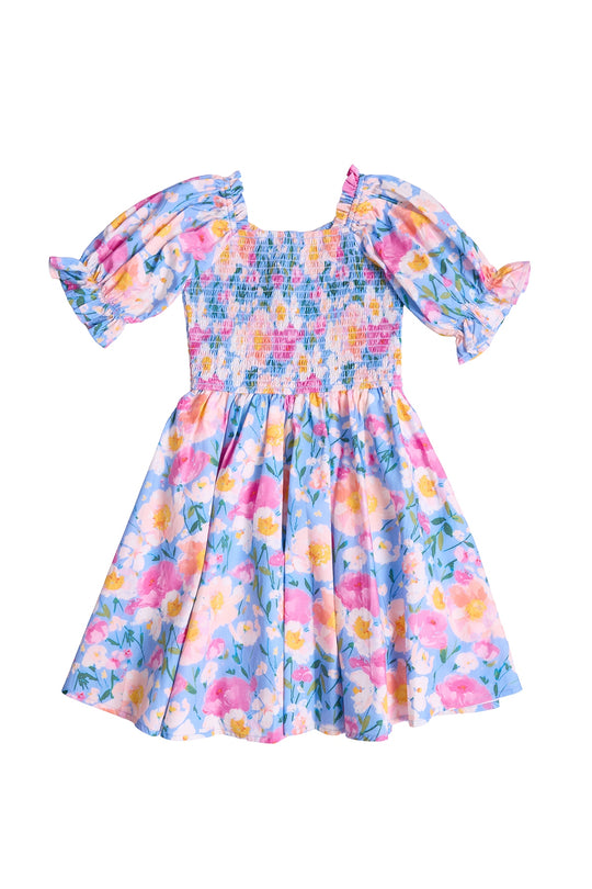 The Mini Teagan Dress in Delia Blue by Ivy City Co is a girls’ cotton dress with puff sleeves, shirred bodice, and flared skirt featuring vibrant pink, yellow, and white floral patterns on a light blue background.