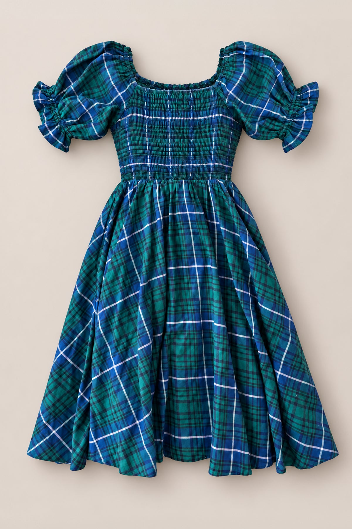The Mini Teagan Dress in Emerald Plaid by Ivy City Co is showcased laid flat on a neutral background, featuring a green, blue, and white plaid pattern with puffed short sleeves, a smocked bodice, and a flared skirt—making it perfect for holiday portraits or as a charming Christmas Eve dress.