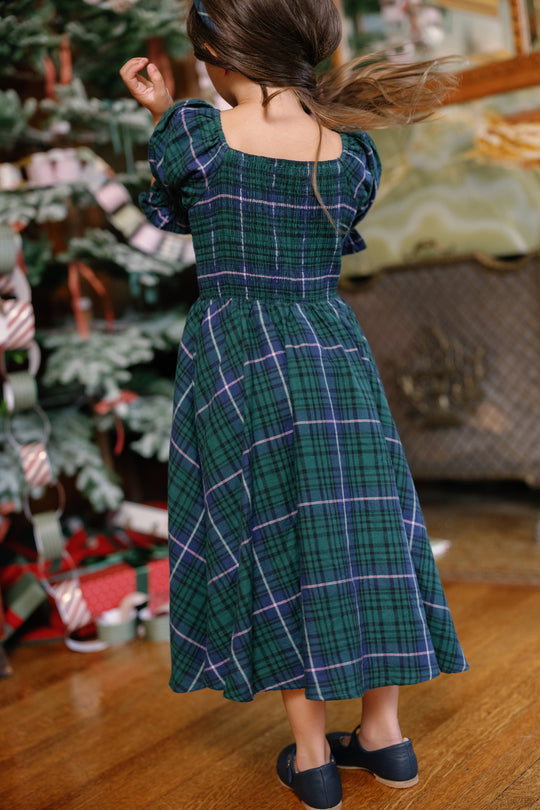 A young girl twirls near a festive Christmas tree, surrounded by presents and paper chains, dressed in the Ivy City Co Mini Teagan Dress in Emerald Plaid—capturing the magic of holiday portraits.