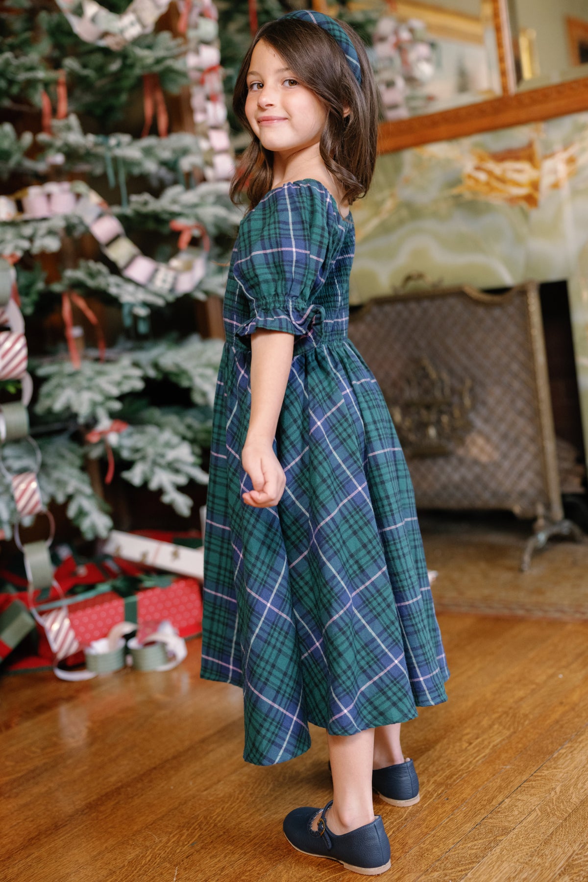 Wearing the Ivy City Co Mini Teagan Dress in Emerald Plaid, a young girl poses indoors by a festively decorated Christmas tree and wrapped presents, smiling over her shoulder—perfectly capturing the joy of holiday portraits.