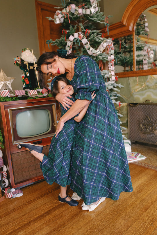 A woman and a girl wearing matching Ivy City Co Mini Teagan Dresses in Emerald Plaid hug and smile beside a vintage TV and beautifully decorated Christmas tree in a cozy, festive room—perfect for capturing heartwarming holiday portraits.