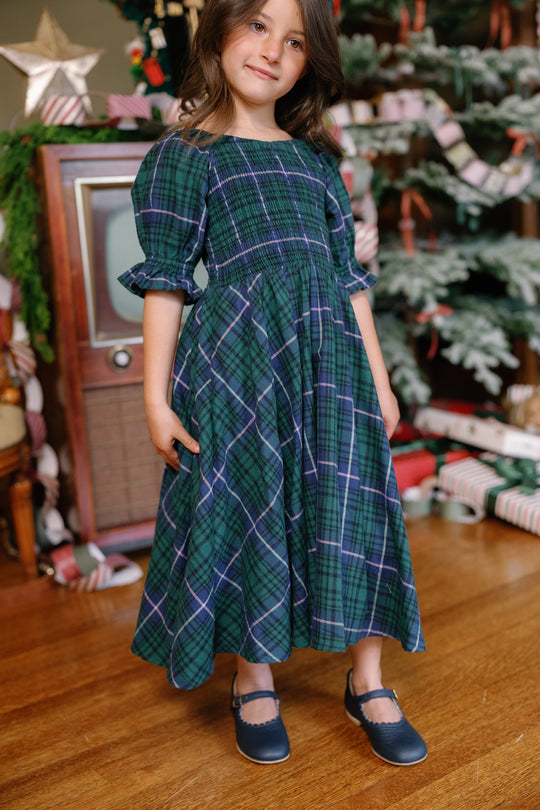 A young girl wearing the Ivy City Co Mini Teagan Dress in Emerald Plaid and blue shoes stands indoors by a vintage TV, Christmas tree, and wrapped presents, smiling gently at the camera—perfect for capturing charming holiday portraits.