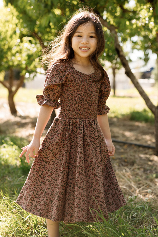 A young girl stands smiling outdoors on a sunny day, surrounded by green trees and grass, wearing the Ivy City Co Mini Teagan Dress in Mauve Woodland Floral with a smocked bodice.