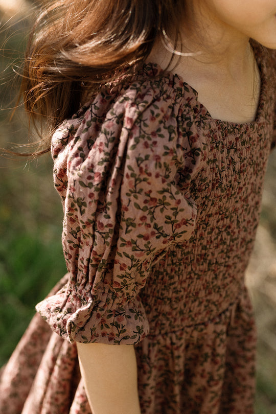 A young child with long brown hair stands outdoors in natural light, wearing the Ivy City Co Mini Teagan Dress in Mauve Woodland Floral—a cotton dress featuring a brown floral pattern, puffed sleeves, and a smocked bodice.