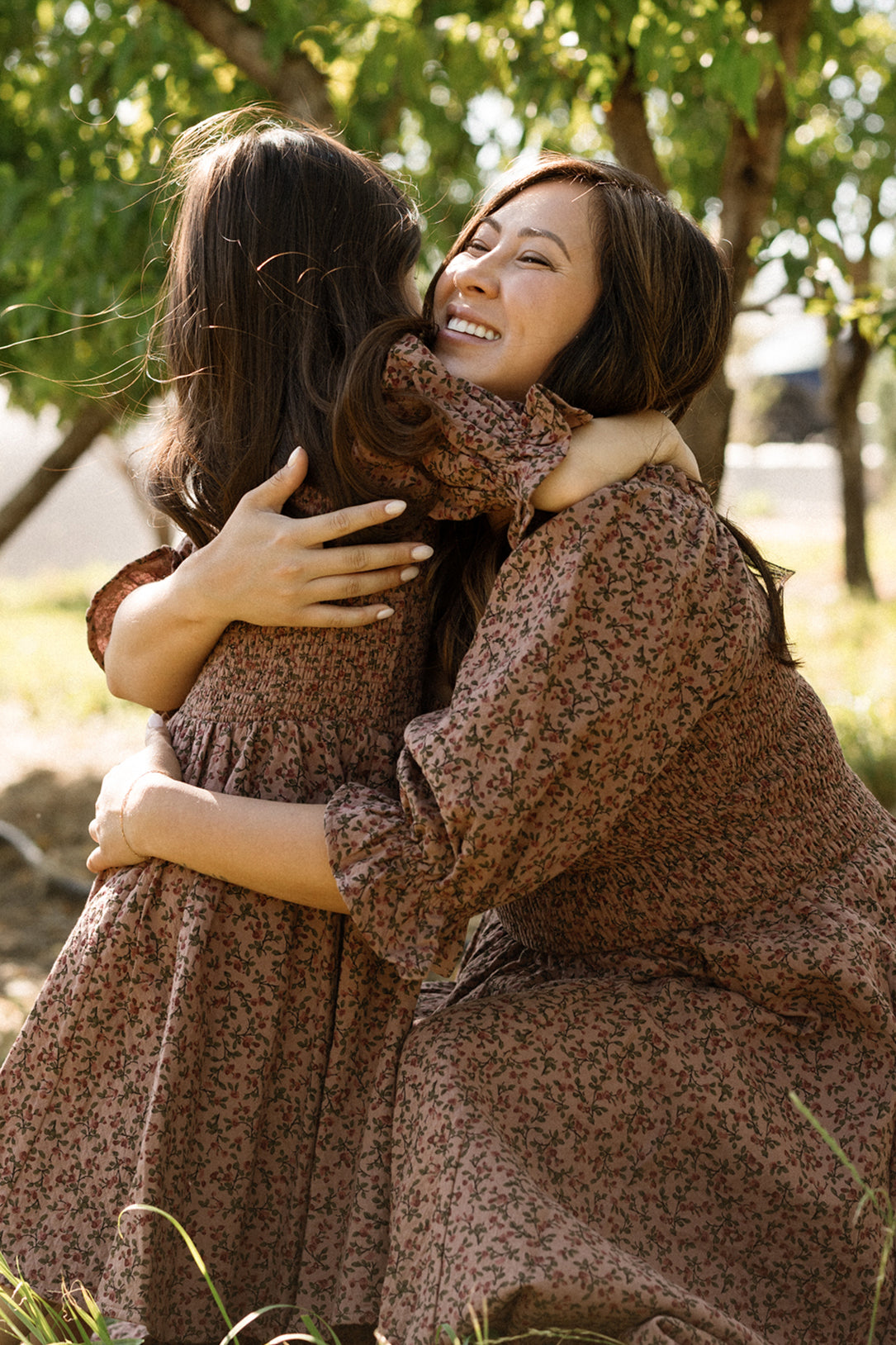 A woman and a young girl, both wearing the Ivy City Co Mini Teagan Dress in Mauve Woodland Floral with charming smocked bodices, hug each other and smile outdoors under leafy trees.