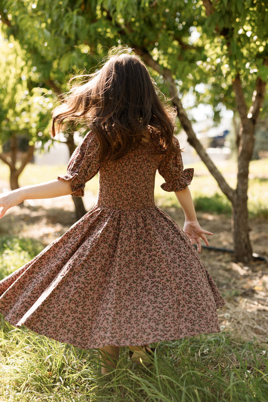 A young girl in the Ivy City Co Mini Teagan Dress in Mauve Woodland Floral twirls joyfully outdoors among green trees, her hair and skirt flowing as she spins, with sunlight filtering through the leaves. The cotton dress’s smocked bodice adds a charming touch to her carefree moment.