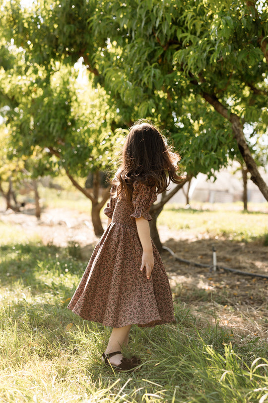 A young girl in the Ivy City Co Mini Teagan Dress in Mauve Woodland Floral stands outdoors among green trees, her long hair flowing as she gazes away from the camera. Sunlight filters through the leaves, casting a warm glow on the grass and illuminating the charming details of her floral cotton dress with its smocked bodice.