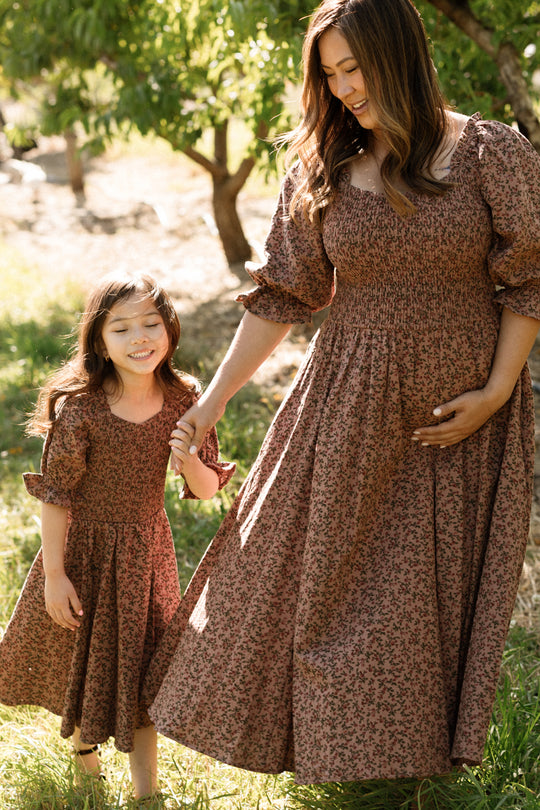 A woman and a young girl smile and hold hands while standing outdoors on sunlit grass, both wearing matching Ivy City Co Mini Teagan Dresses in Mauve Woodland Floral with smocked bodices.