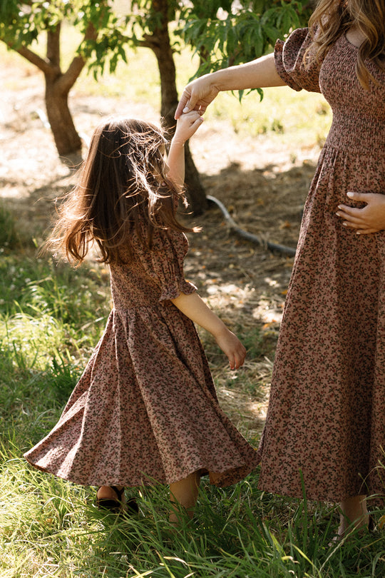 A young girl twirls outdoors among trees and grass, wearing the Ivy City Co Mini Teagan Dress in Mauve Woodland Floral with a smocked bodice, holding hands with an adult in a matching dress.