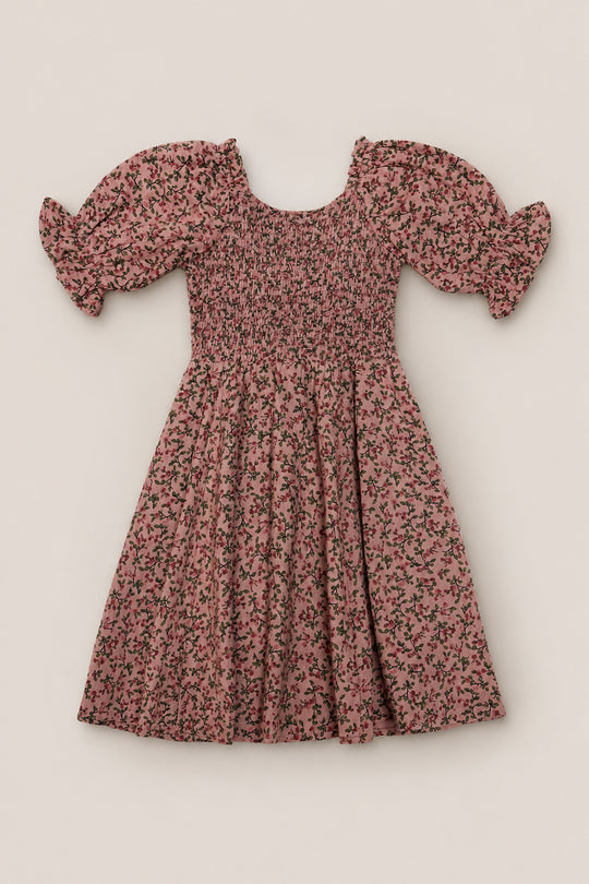The Mini Teagan Dress in Mauve Woodland Floral by Ivy City Co features puffed short sleeves, a smocked bodice, and a flared skirt. This charming cotton dress showcases a delicate black woodland floral pattern throughout and is displayed laid flat on a neutral background.
