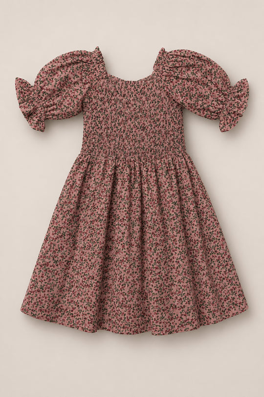 The Mini Teagan Dress in Mauve Woodland Floral by Ivy City Co is a pink cotton dress with short puffed sleeves finished with ruffled ends. Featuring a small floral print, the dress has a square neckline, smocked bodice, gathered waist, and an A-line skirt, all set against a plain light beige background.