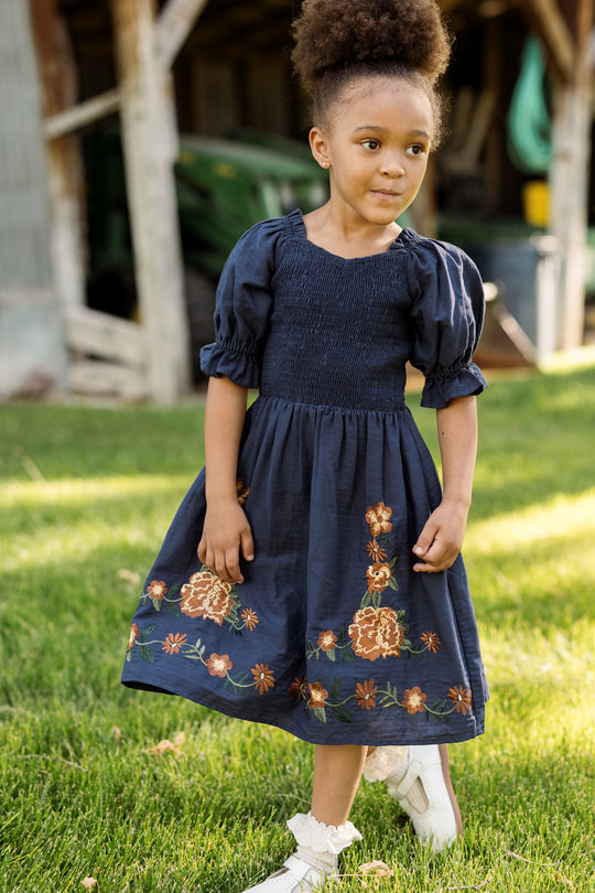 A young girl with a high puff hairstyle wears the Ivy City Co Mini Teagan Dress in Navy Embroidered Fall Floral, paired with white shoes, as she stands on green grass outside near a rustic building.