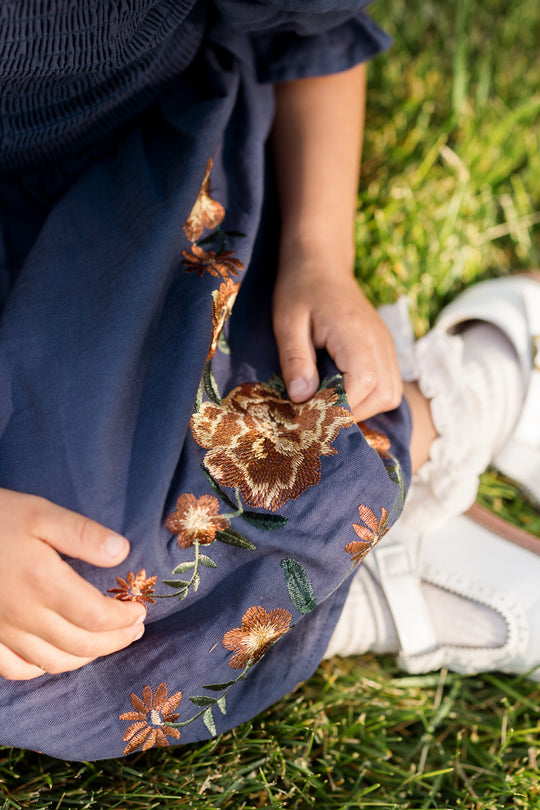 A child wearing the Ivy City Co Mini Teagan Dress in Navy Embroidered Fall Floral, featuring a smocked bodice and delicate floral embroidery, sits on the grass displaying white shoes and socks.