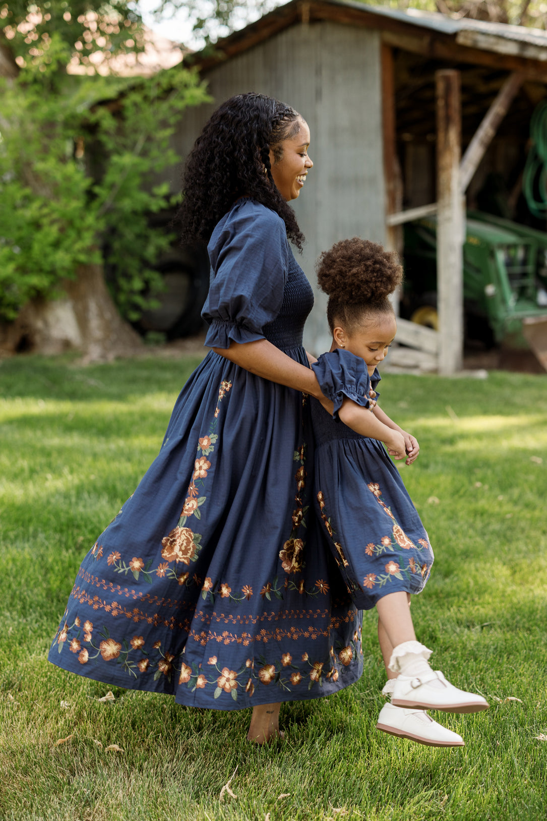 A woman and a young girl, both wearing the Ivy City Co Mini Teagan Dress in Navy Embroidered Fall Floral, hold hands and smile as they walk barefoot on green grass in front of a rustic barn.