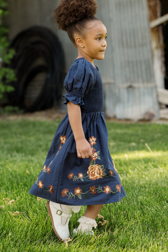 A young girl with curly hair in a bun, wearing the Ivy City Co Mini Teagan Dress in Navy Embroidered Fall Floral and white shoes, walks on green grass beside a corrugated metal building.