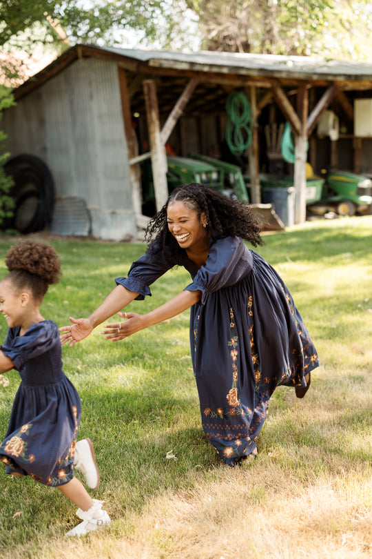 A woman and a young girl, both in matching blue cotton dresses with smocked bodices, laugh and run together on grass near a rustic shed with farm tools in the background. The girl’s outfit is the adorable Ivy City Co Mini Teagan Dress in Navy Embroidered Fall Floral, perfectly complementing their joyful moment.