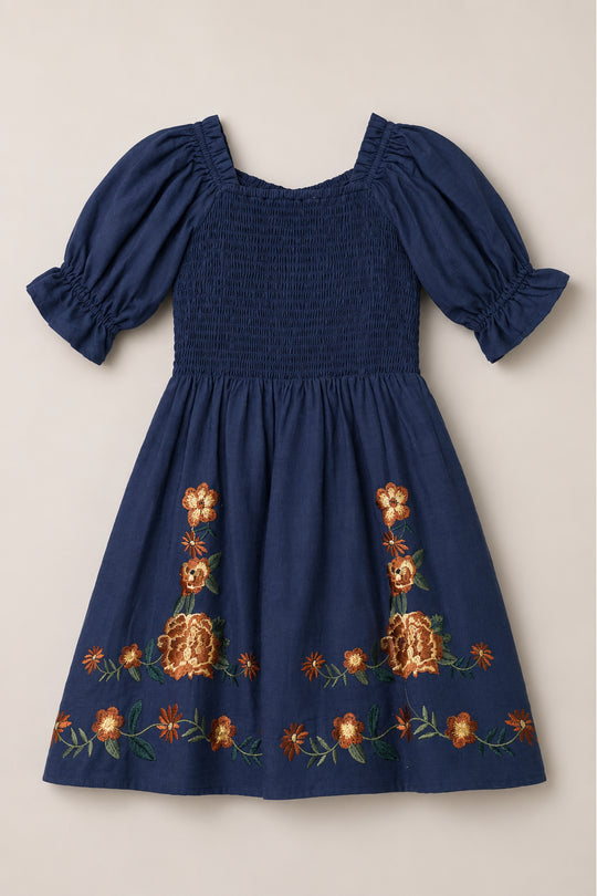 The Mini Teagan Dress in Navy Embroidered Fall Floral by Ivy City Co is laid flat on a neutral background. This short-sleeved navy cotton dress features puffed sleeves, a smocked bodice, and orange and yellow floral embroidery along the skirt.