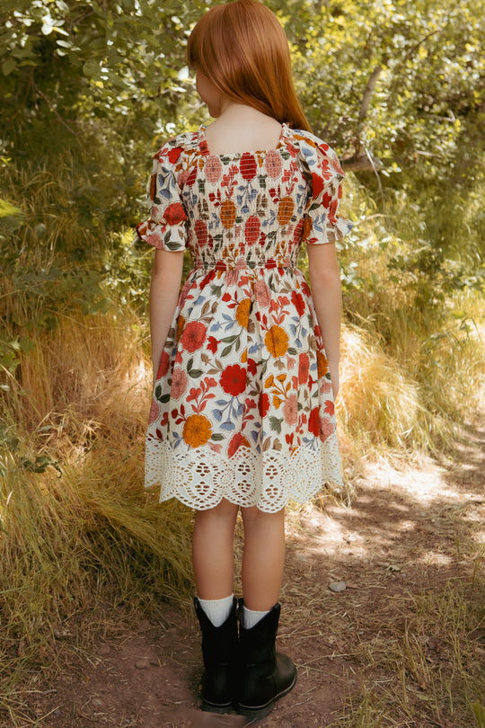 A young girl with long red hair stands on a dirt path surrounded by sunlit greenery. She wears the Ivy City Co Mini Teagan Dress in Sepia Flower, white socks, and black boots, facing away as sunlight filters through the trees.