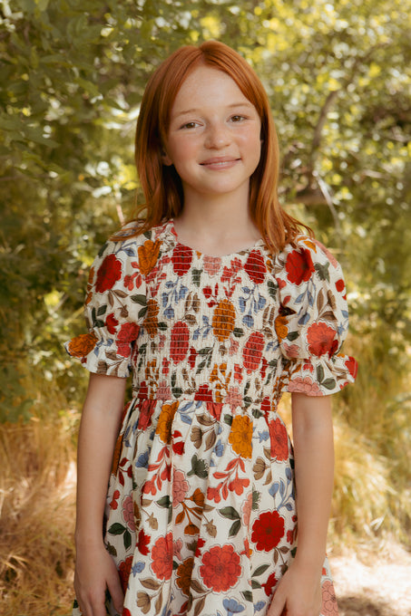 A young girl with red hair smiles outdoors in the Ivy City Co Mini Teagan Dress in Sepia Flower, crafted from 100% cotton with colorful floral patterns, surrounded by green trees and sunlight.