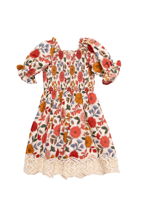 The Mini Teagan Dress in Sepia Flower by Ivy City Co is a girls’ 100% cotton dress featuring puffed short sleeves, smocked bodice, flared skirt, scalloped lace hem, and vibrant red, orange, and yellow floral prints.