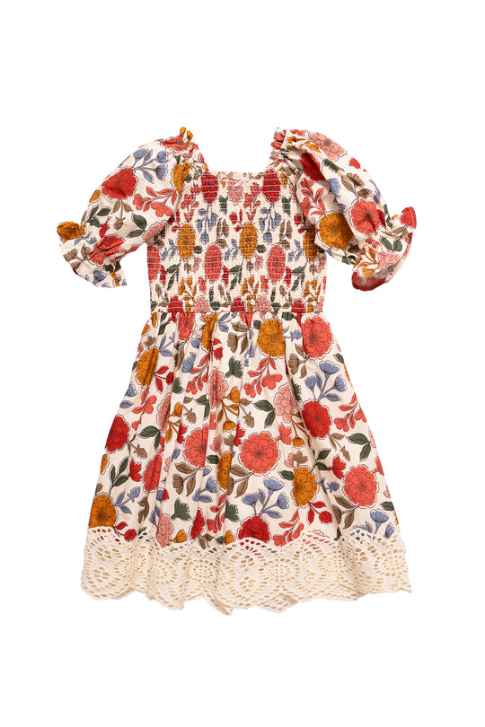 The Mini Teagan Dress in Sepia Flower by Ivy City Co is a girls’ 100% cotton dress featuring puffed short sleeves, smocked bodice, flared skirt, scalloped lace hem, and vibrant red, orange, and yellow floral prints.