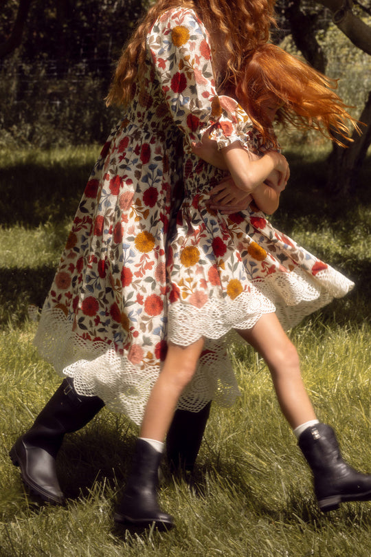 A woman and child with flowing red hair spin on sunlit grass, both wearing Ivy City Co’s Teagan dresses—hers in the Sepia Flower print, her child in the matching Mini Teagan Dress in Sepia Flower—paired with black boots.