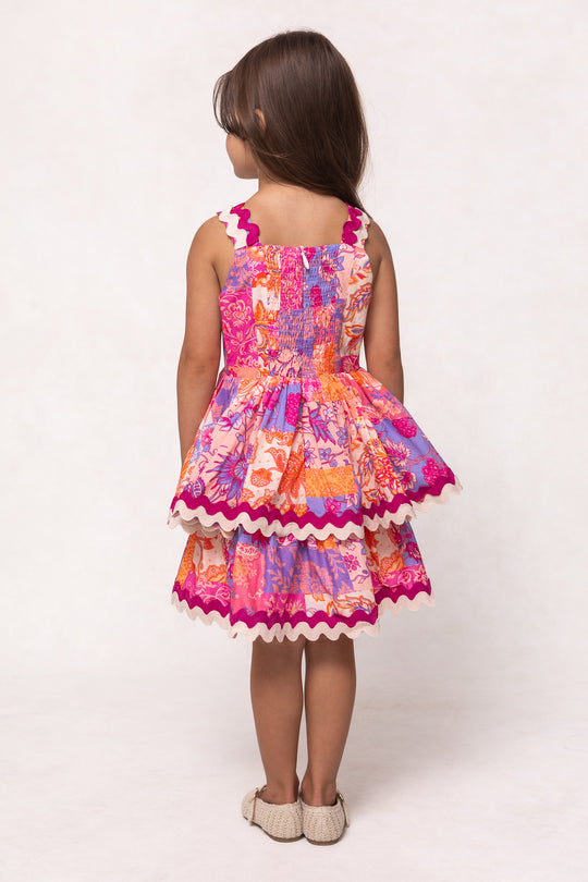 A young girl with long brown hair faces away, wearing the Ivy City Co Mini Tessa Dress in Pink Floral Tile. The knee-length cotton dress features layered ruffles, white wavy trim, and a playful summer vibe, paired with beige shoes.