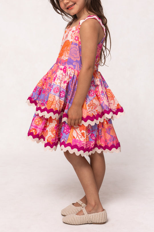 A young girl stands sideways in Ivy City Co’s Mini Tessa Dress in Pink Floral Tile, a sleeveless knee-length cotton dress with orange and purple tiered layers, paired with beige textured shoes, set against a light plain background.