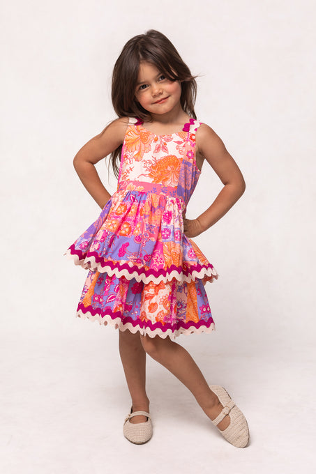A young girl with long brown hair smiles brightly, hands on hips, wearing Ivy City Co’s Mini Tessa Dress in Pink Floral Tile—a sleeveless, knee-length cotton dress with a layered skirt and scalloped trim—against a plain background.