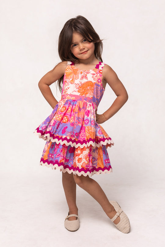 A young girl with long brown hair smiles brightly, hands on hips, wearing Ivy City Co’s Mini Tessa Dress in Pink Floral Tile—a sleeveless, knee-length cotton dress with a layered skirt and scalloped trim—against a plain background.