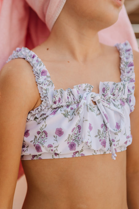 A child wears the Ivy City Co Mini Thea Top in Maeve Floral, featuring purple flowers and green leaves, standing in soft sunlight with a pink towel wrapped around their head.