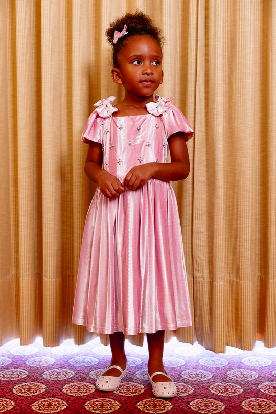 Young girl in a pink dress standing in front of a beige curtain.