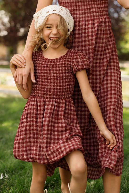 A young girl in the Ivy City Co Mini Tori Dress in Cranberry Popcorn Plaid, styled with puff sleeves and a white knit hat, smiles as she holds an adult’s hand. The adult stands beside her in a coordinating plaid dress with a smocked bodice. They are pictured together on a grassy lawn.