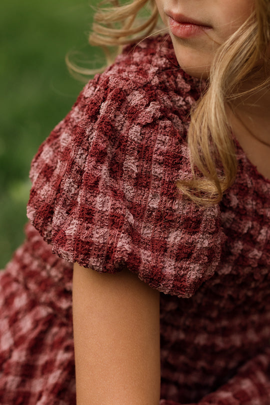 A close-up shot of a person with blonde hair wearing the Ivy City Co Mini Tori Dress in Cranberry Popcorn Plaid, showcasing its maroon and pink plaid textured fabric with puff sleeves. The focus is on the smocked bodice and sleeve details, set against a softly blurred green background.