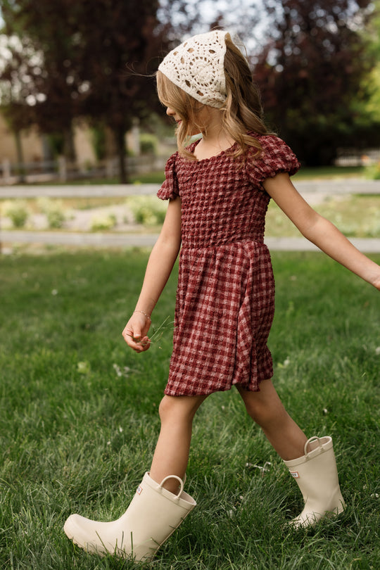 A young girl wearing the Ivy City Co Mini Tori Dress in Cranberry Popcorn Plaid, styled with puff sleeves and a smocked bodice, pairs it with white boots and a cream knit headscarf as she walks on green grass outdoors, looking to the side.