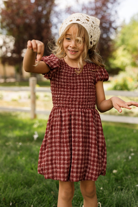 A young girl in the Ivy City Co Mini Tori Dress in Cranberry Popcorn Plaid, featuring puff sleeves, smiles and plays outside on a sunny day while wearing a white crochet hat. She stands on green grass with trees and a wooden fence in the background.