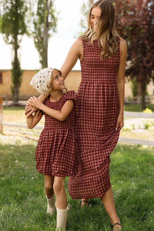 A woman and a young girl smile and walk hand-in-hand on green grass, both dressed in the Ivy City Co Mini Tori Dress in Cranberry Popcorn Plaid featuring puff sleeves and smocked bodices. The girl pairs her dress with cream boots and a crocheted hat. Trees and a building are visible in the background.