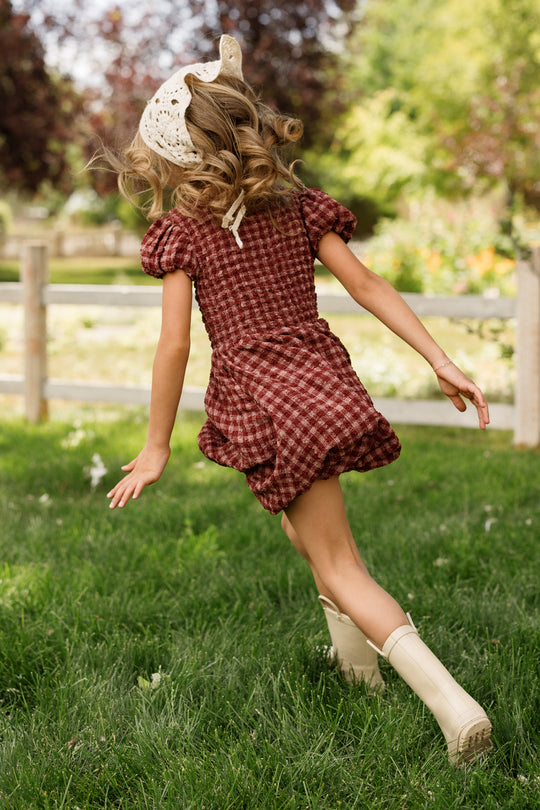 A young girl with curly hair and a cream lace headscarf runs on green grass near a wooden fence and trees, her back to the camera, wearing the Ivy City Co Mini Tori Dress in Cranberry Popcorn Plaid with a smocked bodice and white boots.