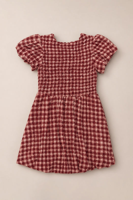 Laid flat on a neutral background, the Ivy City Co Mini Tori Dress in Cranberry Popcorn Plaid features short puff sleeves, a smocked bodice, and a knee-length silhouette adorned with a maroon and white checkered pattern.