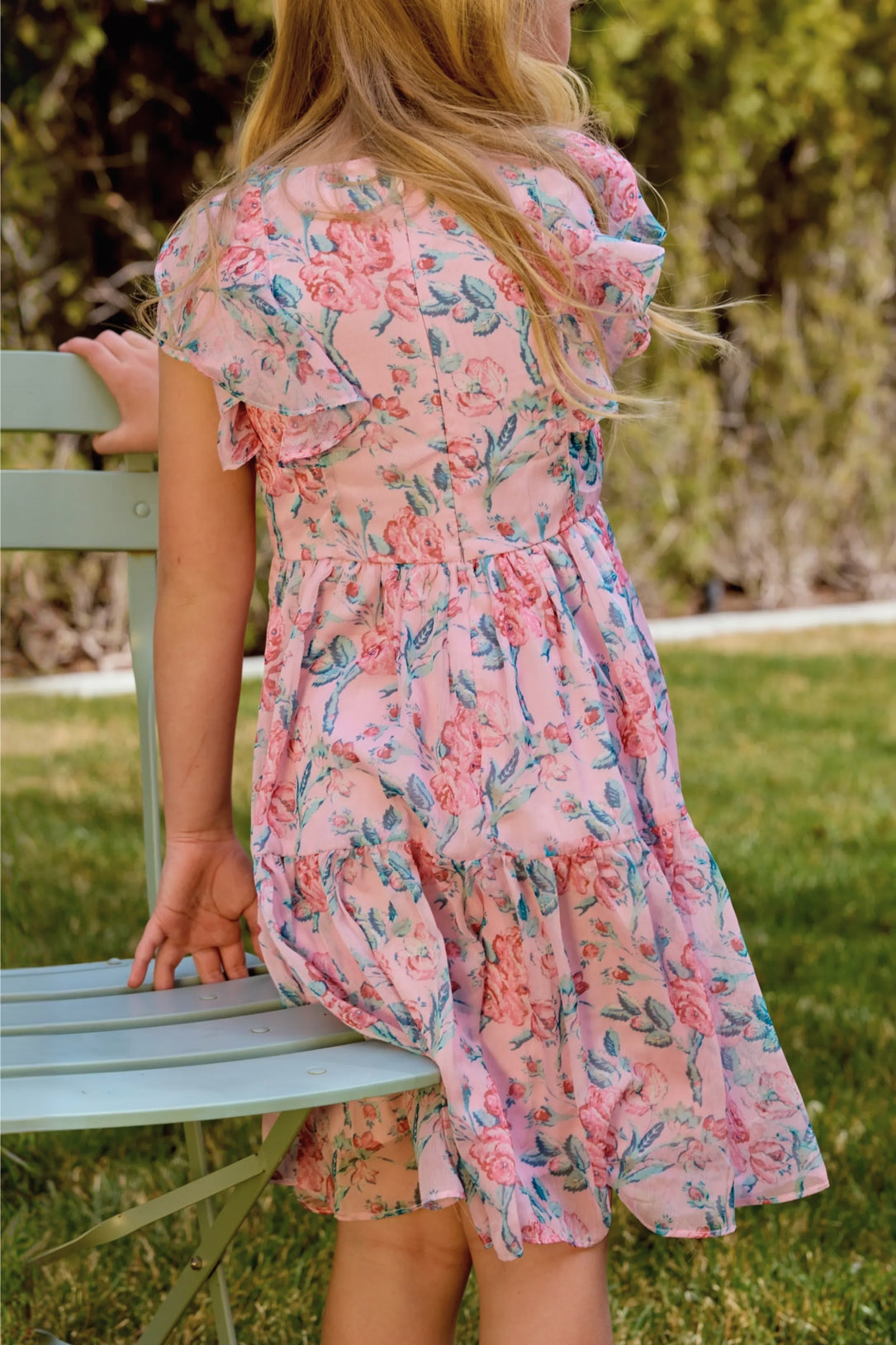 A young girl with long blonde hair wears the Ivy City Co Mini Trixie Dress in Pink Sketched Flower Chiffon as she sits sideways on a light green metal chair outside, surrounded by sunny grass and blurred greenery.