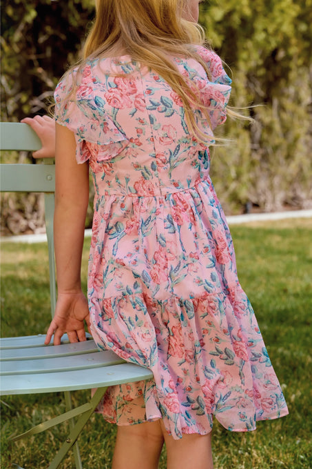 A young girl with long blonde hair wears the Ivy City Co Mini Trixie Dress in Pink Sketched Flower Chiffon as she sits sideways on a light green metal chair outside, surrounded by sunny grass and blurred greenery.