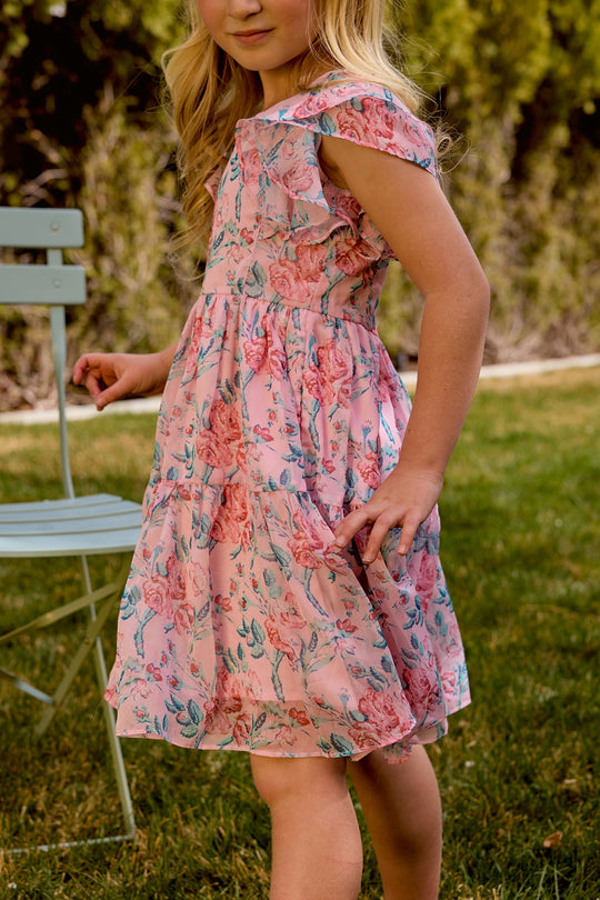 A young girl wears the Mini Trixie Dress in Pink Sketched Flower Chiffon by Ivy City Co, standing outdoors near a light blue chair with greenery in the background—ideal for girls’ summer style or mom and me matching outfits.