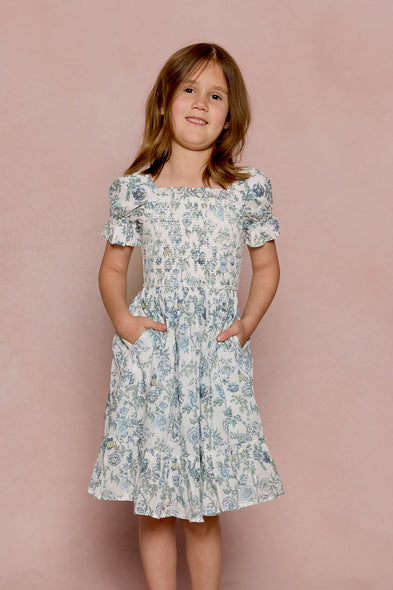 A young girl with shoulder-length brown hair smiles in the Ivy City Co Mini Valencia Dress in Ivory Climbing Vine Bloom, featuring short sleeves and pockets, against a soft pink background.