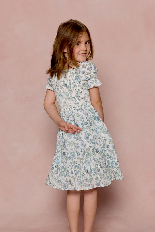 A young girl with light brown hair smiles over her shoulder, wearing the Ivy City Co Mini Valencia Dress in Ivory Climbing Vine Bloom, standing in front of a soft pink background.