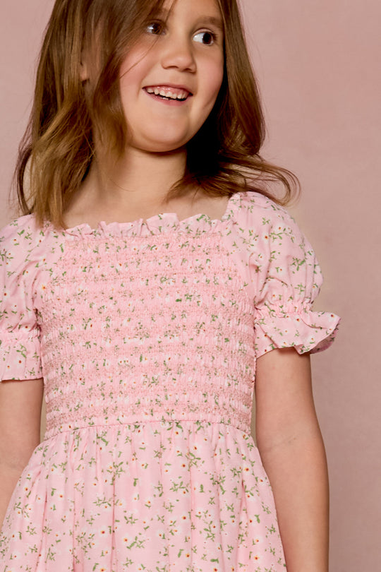 A young girl smiles in the Ivy City Co Mini Valencia Dress in Pink Ditsy Floral, featuring a smocked bodice and short sleeves, as she stands before a matching pink background.