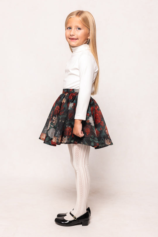 Young girl wearing a white long-sleeve top and floral skirt on a plain background