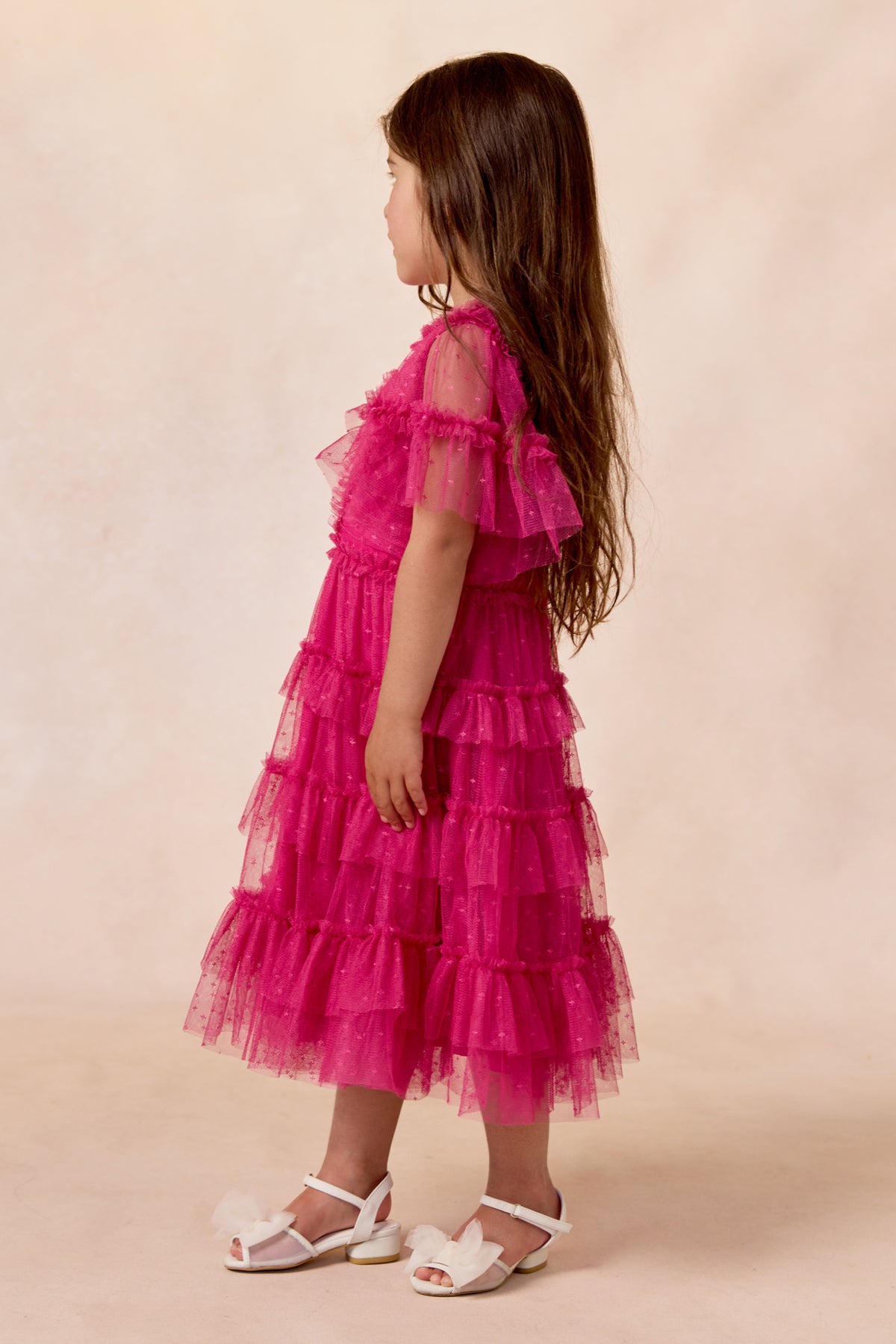 A young girl with long brown hair, wearing the Ivy City Co Mini Whimsical Dress in Fuchsia and white sandals, stands sideways against a light beige background.