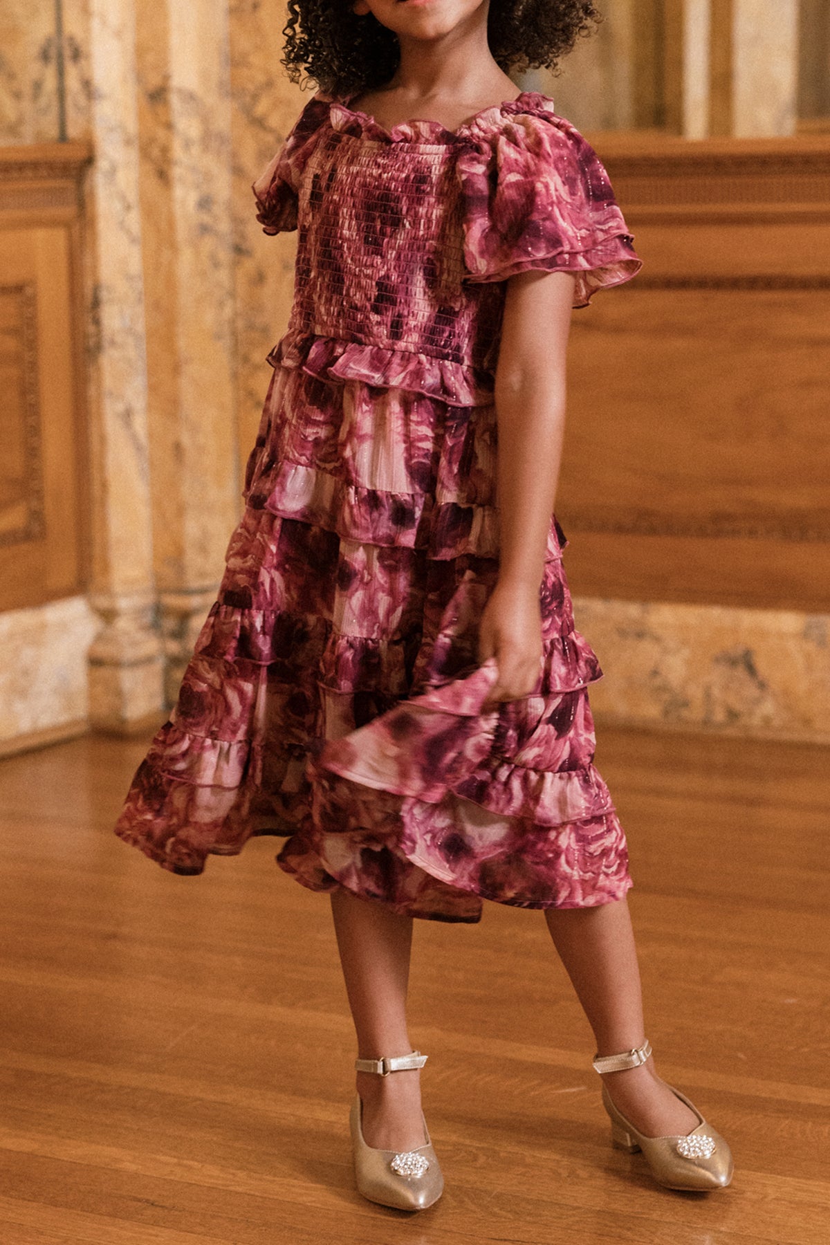 A young child stands indoors on a wooden floor, wearing the Ivy City Co Mini Winnie Dress in Dreamy Roses and shiny gold shoes. She holds part of her skirt, with her face partially out of frame.