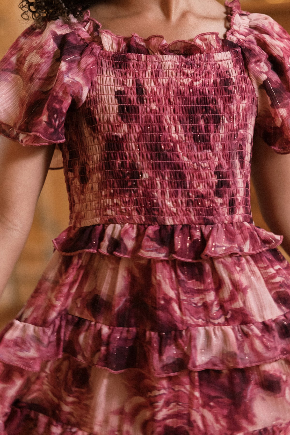 A person wearing the Mini Winnie Dress in Dreamy Roses by Ivy City Co, a girls’ smocked dress featuring pink and burgundy hues with puffed sleeves and ruffled layers. The fabric displays a tie-dye, watercolor-inspired pattern, making it an ideal unique holiday party dress. The image is cropped to show only the midsection of the outfit.